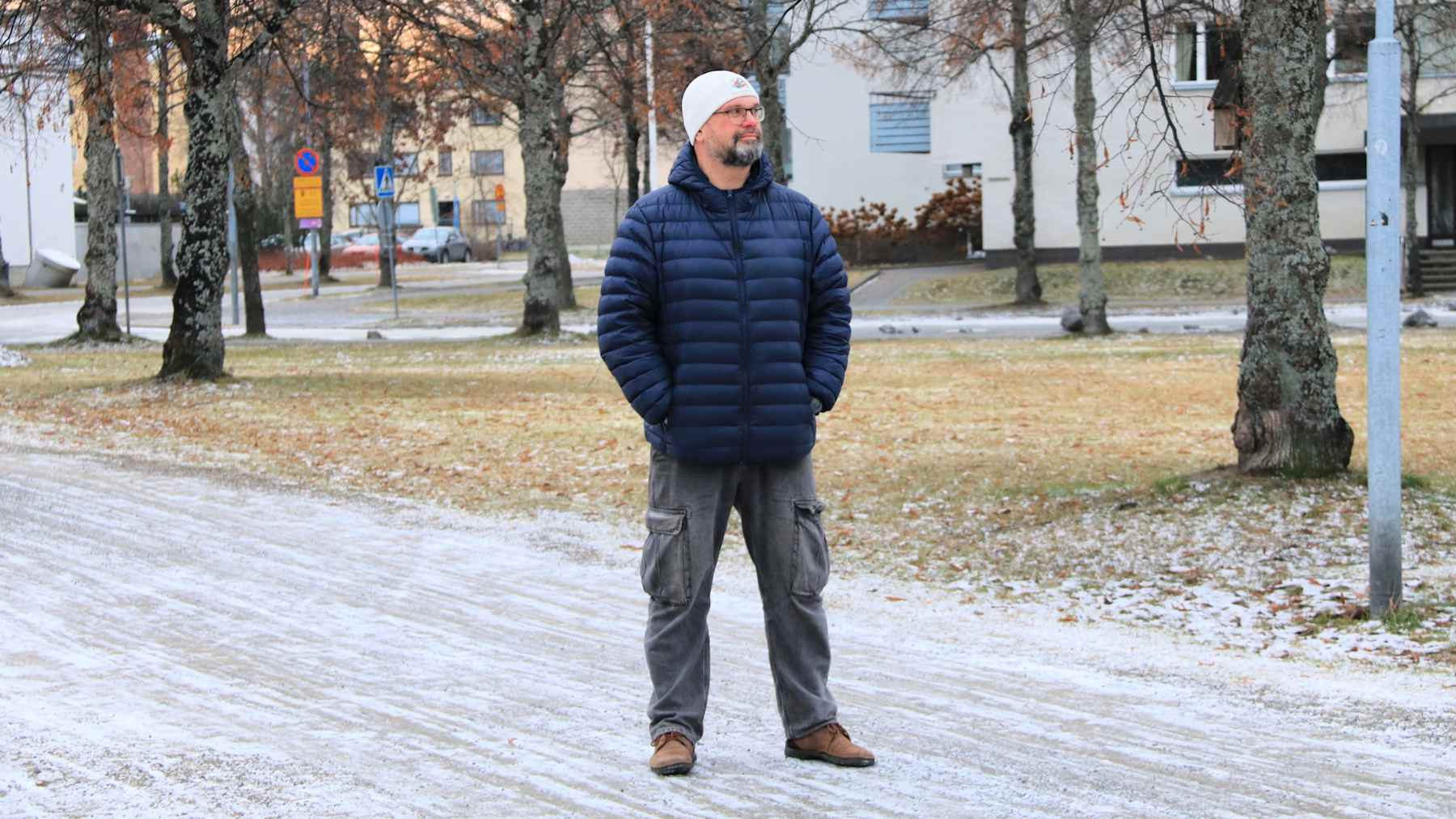 Silmälasipäinen parrakas mies seisoo talvivaatteissa puistossa. Hänellä on kädet taskuissa.