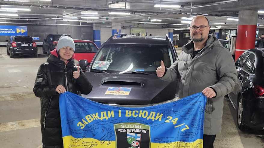 Mies ja nainen seisovat parkkihallissa auton edessä. Heillä on käsissään Ukrainan lippu, johon on kirjoitettu mustalla tussilla tekstiä.