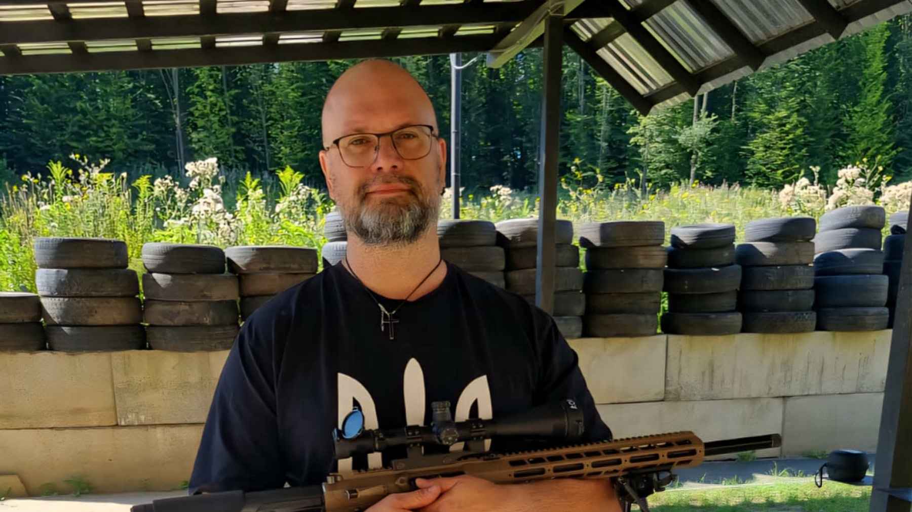 Silmälasipäinen ja parrakas mies pitelee käsissään konetuliasetta.