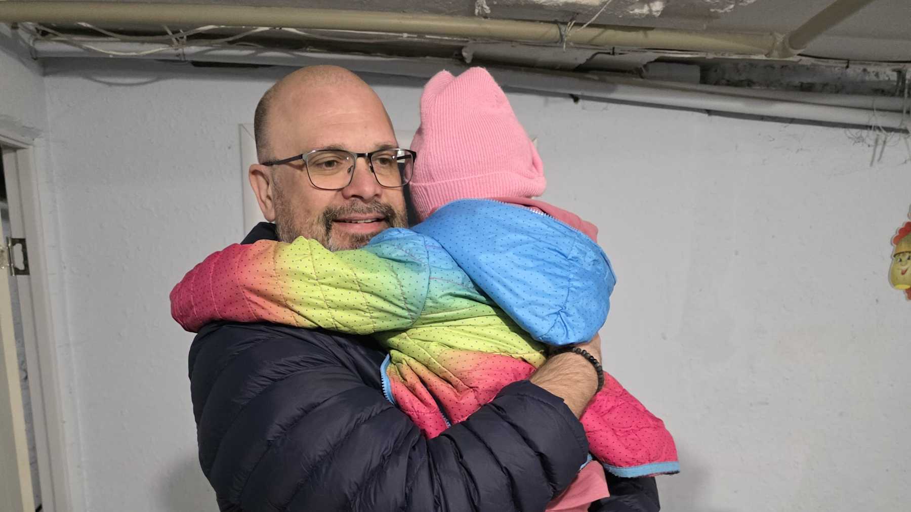 Silmälasipäinen ja parras mies halaa värikkääseen ulkotakkiin pukeutunutta lasta.