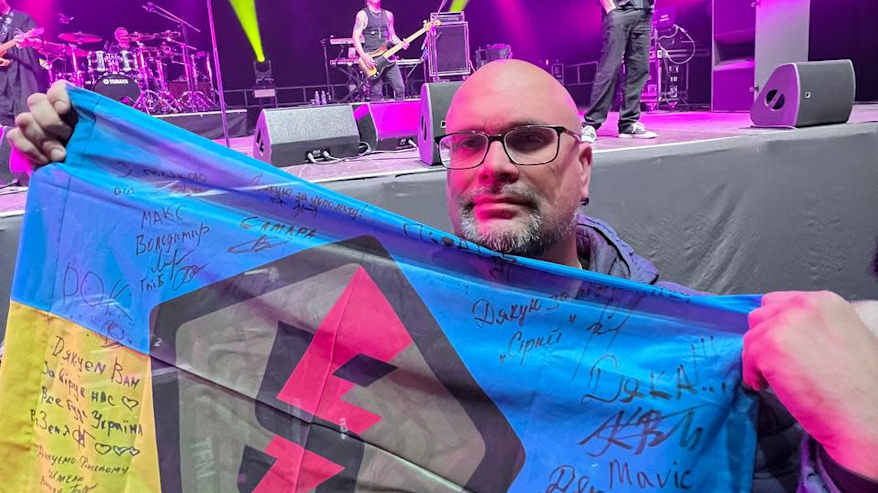 Silmälasipäinen mies poseeraa musiikkikonsertissa Ukrainan lipun kanssa.