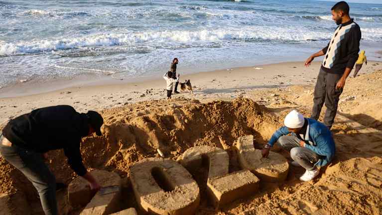 Kolme miestä muotoilee hiekkarannalla hiekasta vuosiluvun 2026.