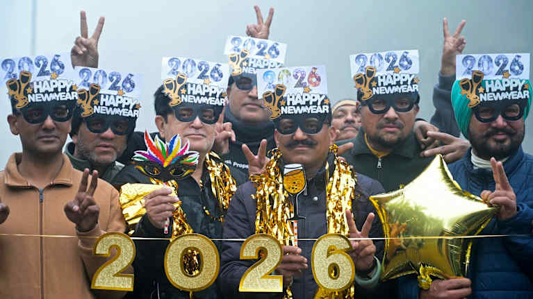 Joukko miehiä pukeutuneena silmänaamioihin ja glitteriin, edessään kyltti, jossa lukee 2026.
