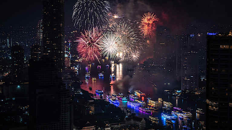 Näkymä Bangkokin ilotulitteiden valaisemaan kaupungin keskustaan, jossa joella on erilaisia laivoja.