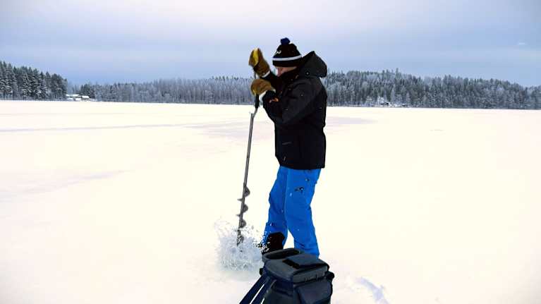 Mies seisoo lumisella järven jäällä ja kairaa jäähän reikää.