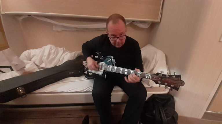 Edesmennyt toimittaja Mikko Tuomikoski istuu sängyllä ja pitelee sähkökitaraa.
