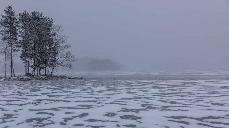 Saari kaukaisuudessa talvipäivänä. Sankka lumisade heikentää etäisyyttä. Etualalalla järven jäätynyt pinta, johon on kinostunut lunta.
