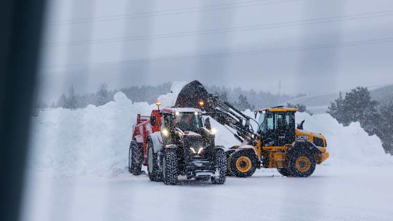 Keltainen traktori lastaa lunta toisen traktorin peräkärryyn. Taustalla on iso lumikasa, taivaalta pyryttää sakeasti lunta.