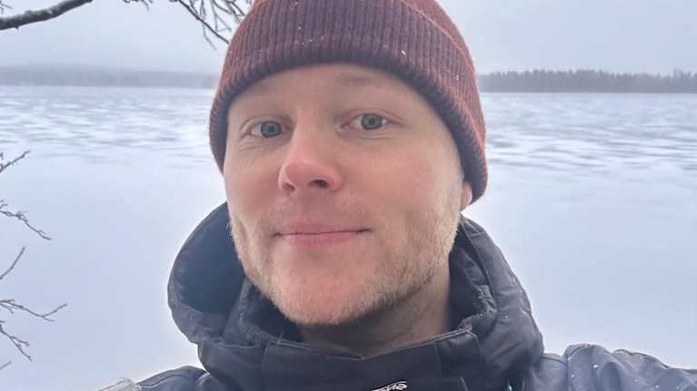 Mies on talvivaatteissa ulkona. Taustalla näkyy jäätynyt järvi ja puun oksia.