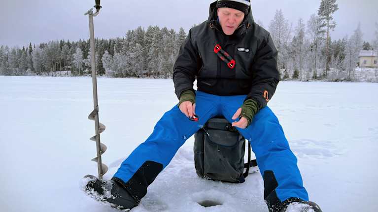 Mies istuu lumisella järven jäällä ja pilkkii.