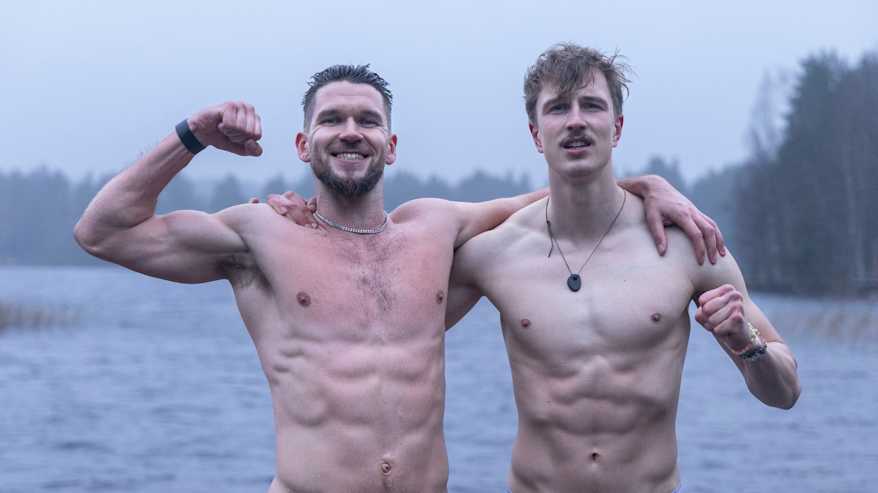 Kaksi paidatonta miestä seisoo lehdettömän metsä- ja järvimaiseman edessä ilman paitaa kädet toistensa hartioilla.