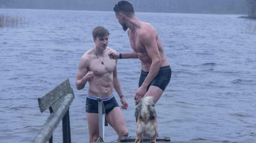 Kaksi miestä nousee laiturille syksyisestä järvestä ja on juuri iskemässä nyrkkejään yhteen. Laiturilla on myös koira.