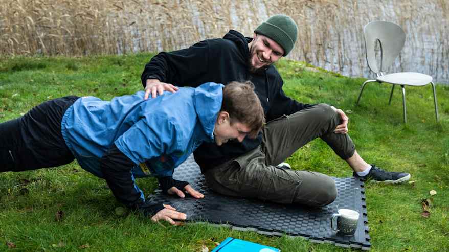 Kaksi nauravaa miestä nurmikolla. Toinen miehistä punnertaa ja toinen istuu vieressä pitäen kättään punnertavan miehen selällä.