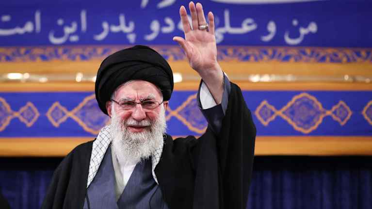 Turbaanipäinen vanha mies käsi pystyssä tilaisuudessa.