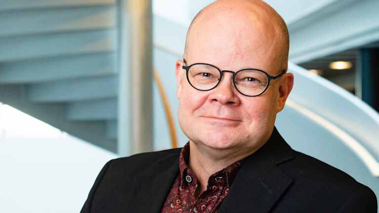 Mies mustassa pikkutakissa katsoo kameraan taustalla kierreportaat.