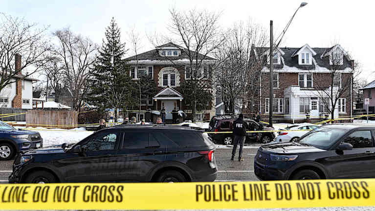 Talojen eteeen on pysäköity autoja, joiden keskellä seisoo FBI:n agentti, alue on eristetty poliisin keltaisella nauhalla.