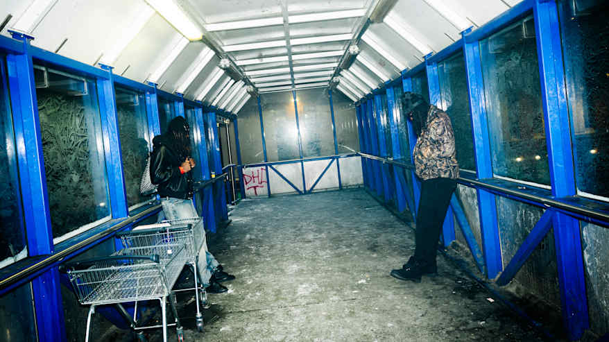 Kaksi henkilöä seisoo graffitien koristamassa kävelytunnelissa, yhden henkilön vieressä ostoskärryt. 