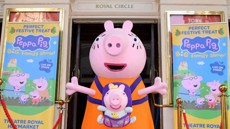 Possuksi pukeutunut hahmo, jolla on pikkupossu kantorepussa, seisoo vaaleanpunaisella matolla oviaukossa.