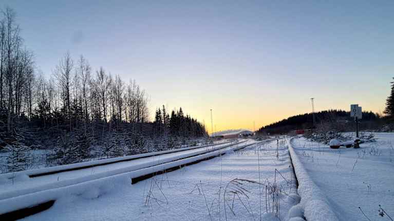 Lumen peittämät junaraiteet, horisontissa kajastaa aurinko.