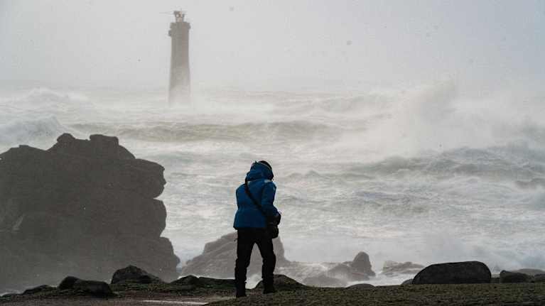 Myrskyisä meri ja majakka taustalla, kun henkilö kävelee kalliot reunustavalla rannikolla.