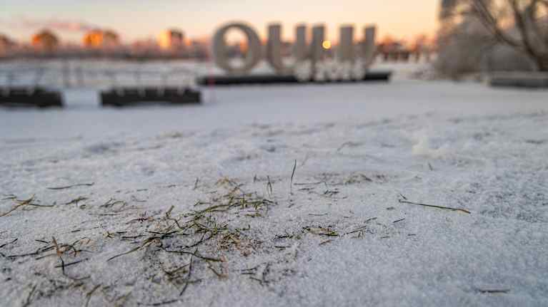 Luminen maisema auringonlaskun aikaan, etualalla näkyy ruohoa lumen peitossa ja taustalla näkyy "Oulu" teos.