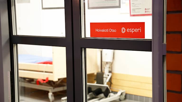Lasiovi, jonka takana näkyy sairaalasänky ja hoitolaitteita. Ovessa on kyltti, jossa lukee Hoivakoti Otso ja Esperi.