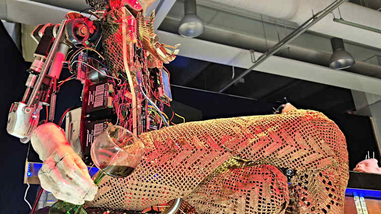 Lähikuva baaritiskin tuolilla istuvasta naisrobotista jolla on viinilasi kädessään.