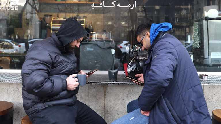 Kaksi miestä istuu ulkona kahvilan penkeillä ja käyttää puhelimia.