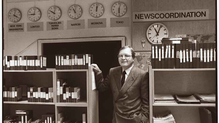  Jaakko Heino kahden kirjahyllyn välissä. Seinällä 6 viisarikelloa, joissa eri maiden pääkaupunkien kellon aika.