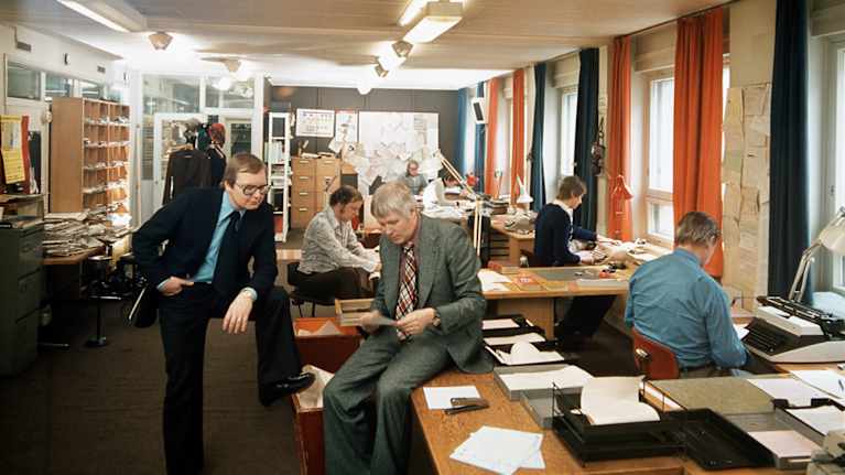 Toimistossa Jaakko Heino nojaa jalkaansa, jonka on nostanut laatikon reunalle ja Aarne Tanninen istuu pöydän reunalla lukemassa paperista.