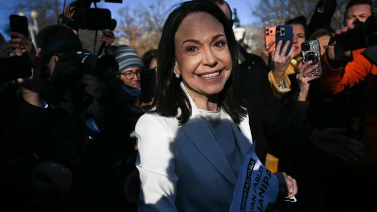 Valkoiseen jakkupukuun pukeutunut nainen hymyilee kameroille ihmisjoukon keskellä.