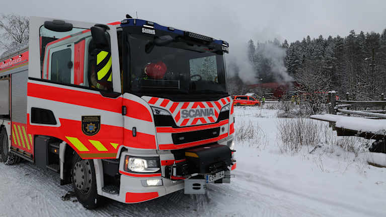 Paloauto talvisessa maisemassa taustalla nousee savua.