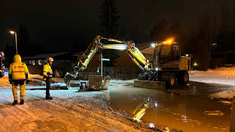 Kaivinkone kaivaa auki katua katuvalojen loisteessa, työmiehet valvovat vieressä.