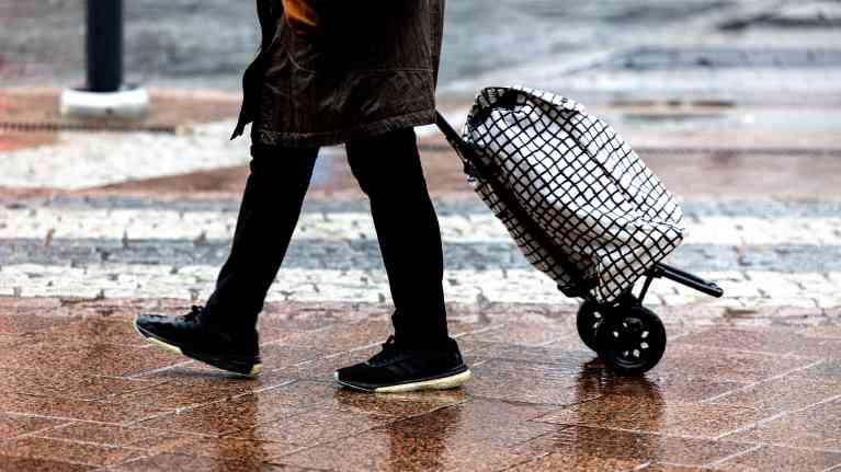 Eläkeläinen vedettävän ostoskassin kanssa suojatien edessä.