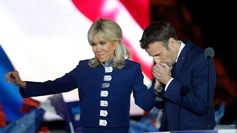 Emmanuel Macron lutar sig över sin fru Brigitte, håller om hennes hand och pussar den. Brigitte vinkar medan hon står på scen.