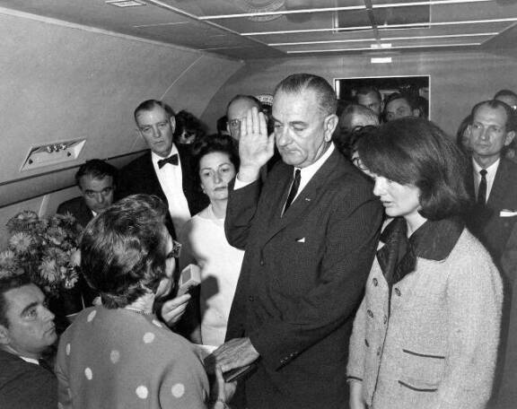 John F. Kennedyn kuolinpäivänä varapresidentti Lyndon B. Johnson vannoi virkavalansa Air Force One -lentokoneen matkustamossa. Hän halusi edeltäjänsä lesken Jacquelinen (kuvassa oikealla) seisovan rinnallaan historiallisena hetkenä. Johnsonin vasemmalla puolella hänen vaimonsa Lady Bird.