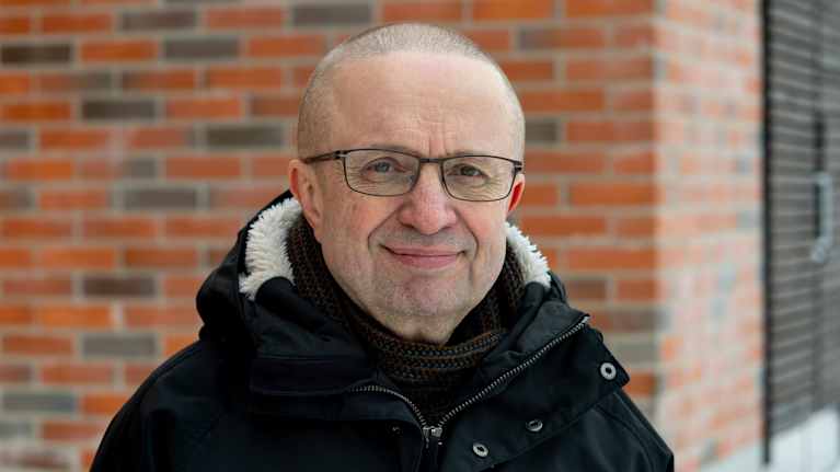 Silmälasipäinen mies jolla on kusta takki, katsoo kohti kameraa.