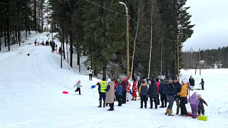 Pulkkamäki metsän keskellä. Lapsi laskee ja ylhäällä toiset odottavat vuoroaan. Alhaalla on katsojia.