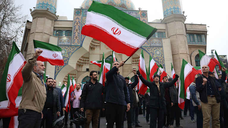 Joukko miehiä seisoo moskeijan edustalla ja heiluttaa Iranin lippuja.