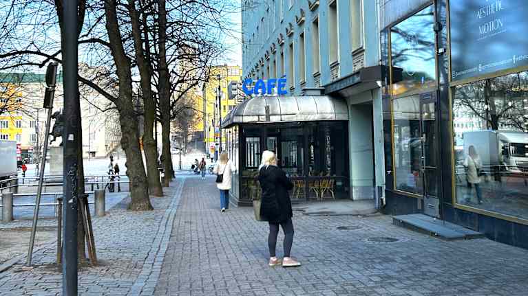 Kuvassa on korkea kerrostalo, jossa on liikerakennus. Katukuvassa näkyy sininen Sinuhe-valomainos ja ihmisiä kadulla.