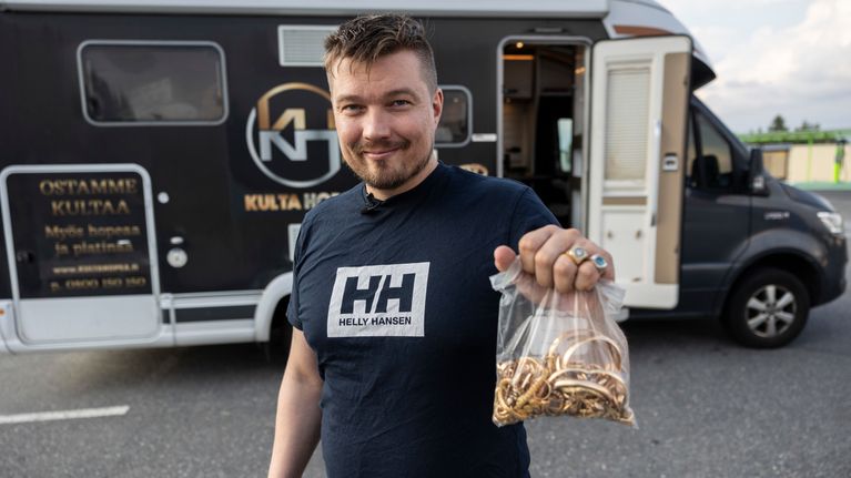Kultakauppias Matias Kerminen pitää kädessään läpinäkyvää muovipussia, jossa kultaisia rannekoruja ja sormuksia. Taustalla mainostekstein teipattu asuntoauto.