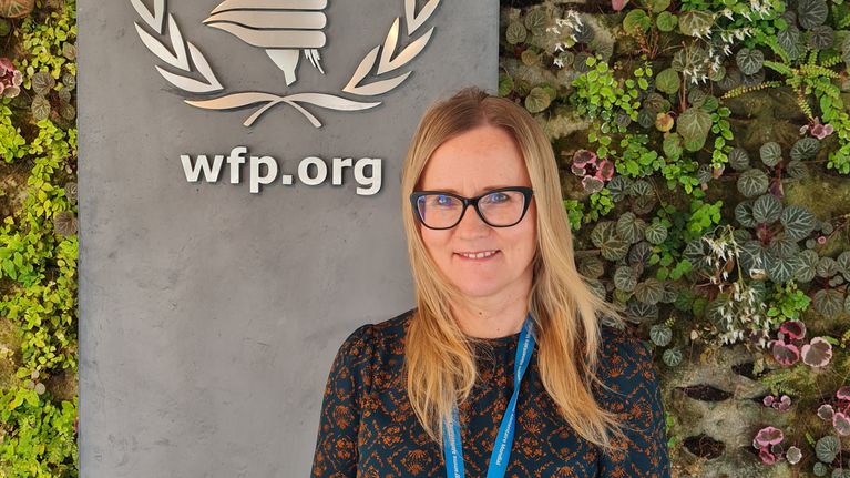 Nainen seisoo kasvot kameraan päin YK:n alaisen Maailman ruokaohjelman WFP:n logon edessä.