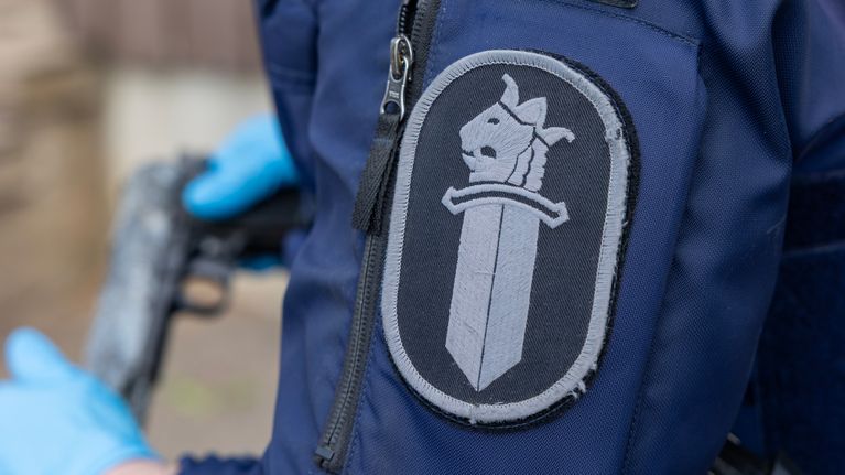 Poliisin hihamerkki, jossa poliisimiekka. Taustalla poliisi pitää kädessään kuulakäsiasetta.