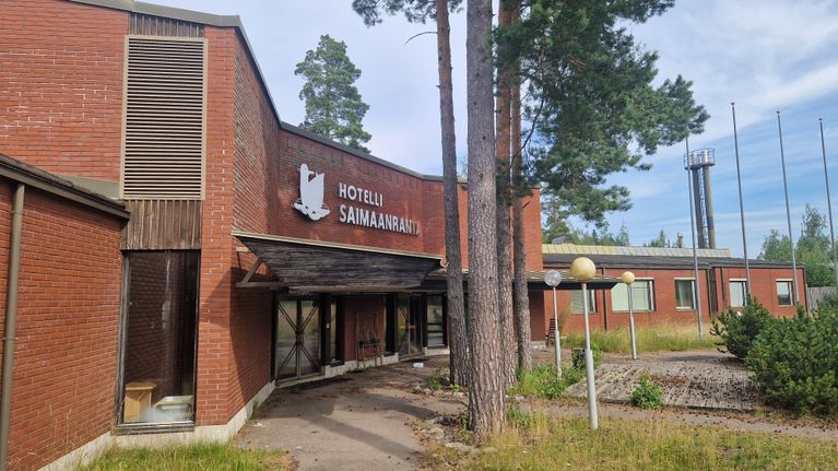 Kuvassa on taipalsaarelaisen hotelli Saimaanrannan sisäänkäynti. Piha-alueella on korkea heinikko, koska hotelli on suljettuna pitkään. 