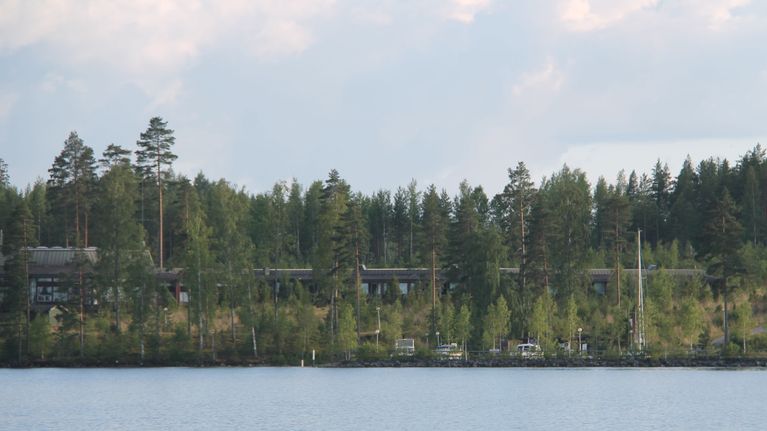 Kuvassa näkyy hotelli Saimaanranta Saimaalta kuvattuna. Hotelli on mäntymetsikon suojassa, etualalla on veneitä