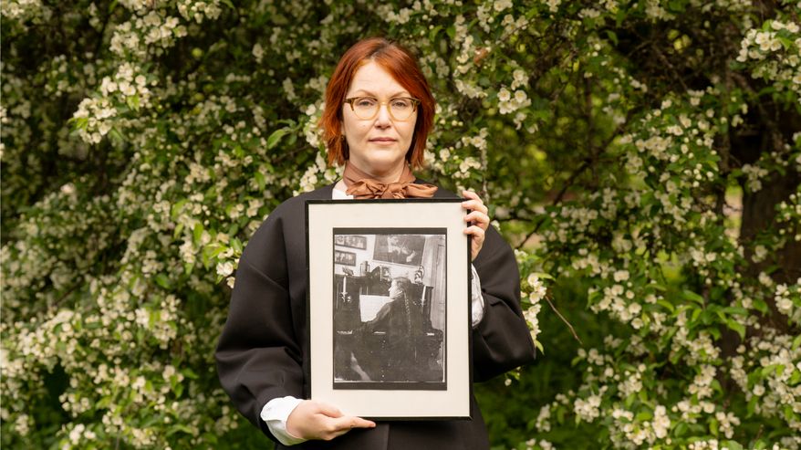 Kuvassa muusikko Astrid Swan seisoo pensaan edessä, katsoo kameraan ja pitää käsissään raamitettua mustavalkovalokuvaa.