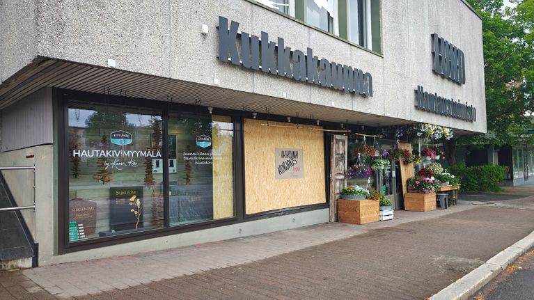 Kukkakaupan julkisivu, jossa yksi näyteikkuna korvattu vanerilla. Vanerissa teksti "Ikkunakorjaus, hyvää kesää!"