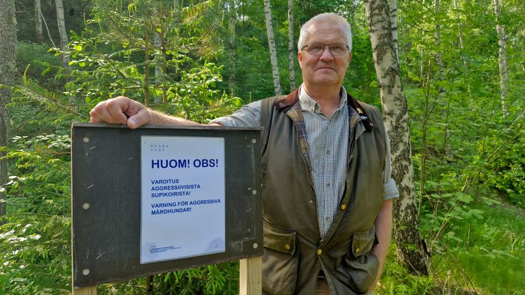 Kauluspaitaan ja vihreään maastoliiviin pukeutunut mies metsässä nojaamassa opaskylttiin.