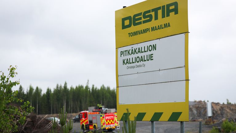 Destian työmaasta kertova kyltti. Taustalla aidan takana näkyy pelastuslaitoksen auto.