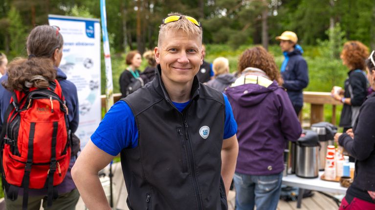 Pyry Rinkinen seisoo terassilla ja katsoo kameraan hymyillen. Takana ihmisiä ja vehreää luontoa.